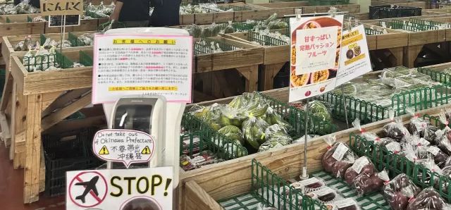 うまんちゅ市場の売り場に立つ、飛行機に斜線を引いたSTOPの札と「不要帯出 沖縄県」の表示