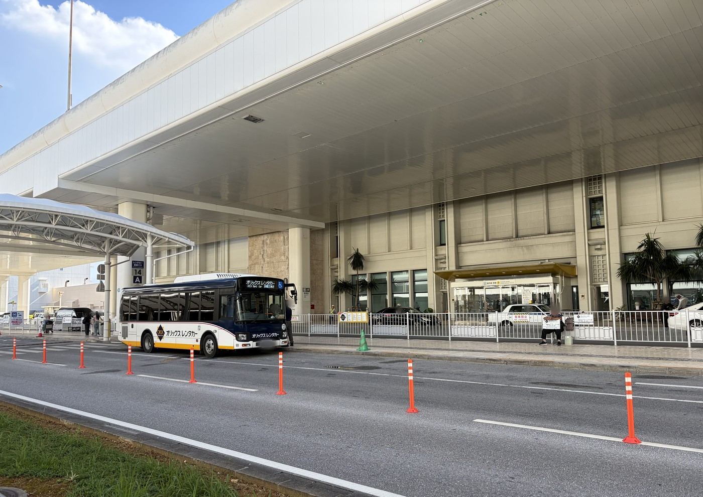 那覇空港の乗り場に停まるレンタカーの送迎バス。歩道ではスタッフが案内板を持って利用者を待っている
